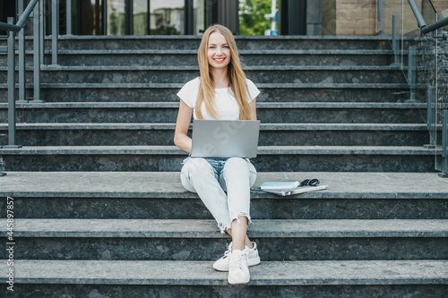 Young student girl sitting with laptop on stairs near university smiling working and look camera