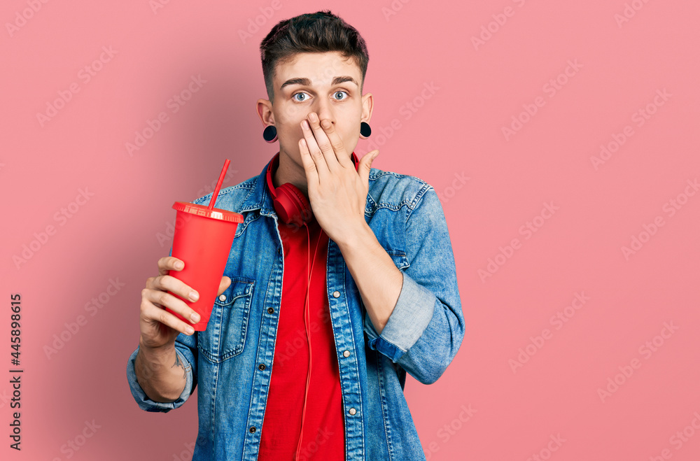 Young caucasian boy with ears dilation drinking glass of cola beverage covering mouth with hand, shocked and afraid for mistake. surprised expression Young caucasian boy with ears dilation drinking glass of cola beverage covering mouth with hand, shocked and afraid for mistake. surprised expression