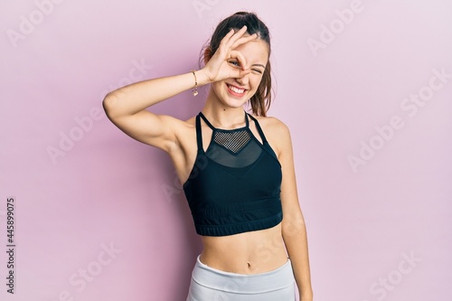 Young brunette woman wearing sportswear smiling happy doing ok sign with hand on eye looking through fingers