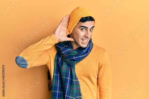 Handsome hispanic man wearing wool hat and winter scarf smiling with hand over ear listening an hearing to rumor or gossip. deafness concept. © Krakenimages.com