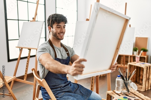 Young hispanic artist man smiling happy holding canvas at art studio.
