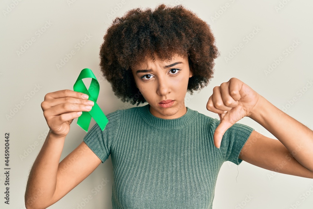 Young hispanic girl holding support green ribbon with angry face, negative sign showing dislike with thumbs down, rejection concept Young hispanic girl holding support green ribbon with angry face, negative sign showing dislike with thumbs down, rejection concept