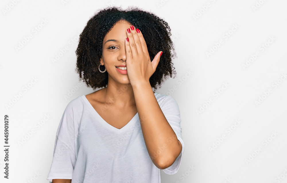 Young african american girl wearing casual clothes covering one eye with hand, confident smile on face and surprise emotion. Young african american girl wearing casual clothes covering one eye with hand, confident smile on face and surprise emotion.