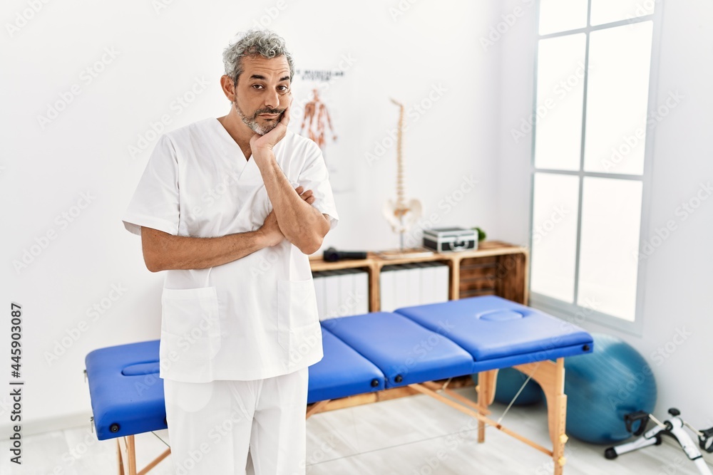 Middle age hispanic therapist man working at pain recovery clinic thinking looking tired and bored with depression problems with crossed arms. Middle age hispanic therapist man working at pain recovery clinic thinking looking tired and bored with depression problems with crossed arms.