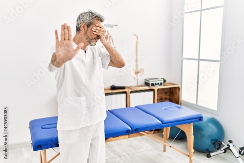 Middle age hispanic therapist man working at pain recovery clinic covering eyes with hands and doing stop gesture with sad and fear expression. embarrassed and negative concept.