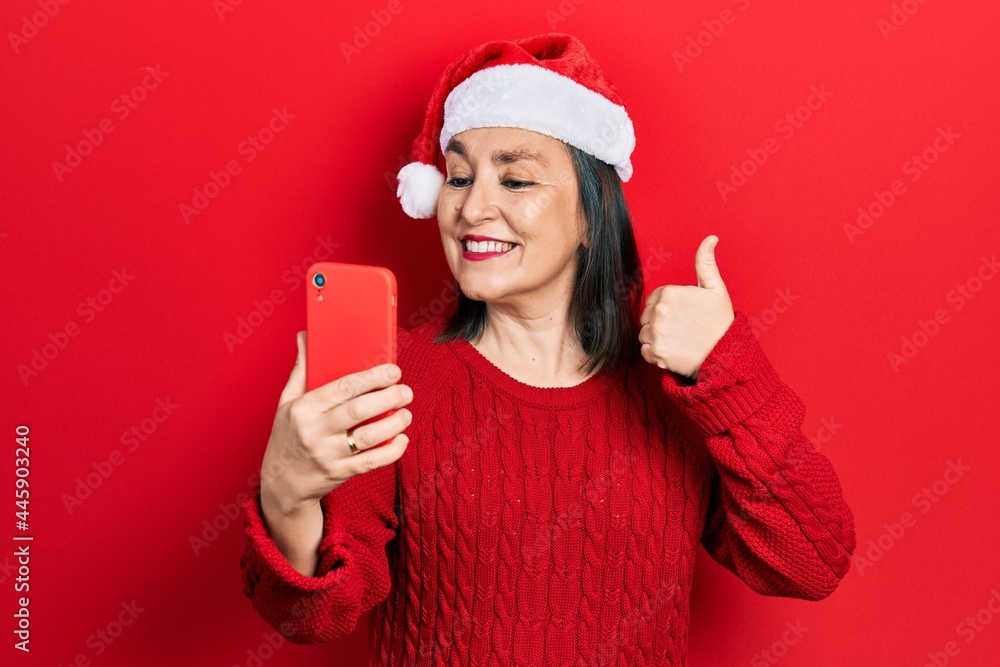 Middle age hispanic woman wearing christmas hat having video call using smartphone smiling happy and positive, thumb up doing excellent and approval sign Middle age hispanic woman wearing christmas hat having video call using smartphone smiling happy and positive, thumb up doing excellent and approval sign