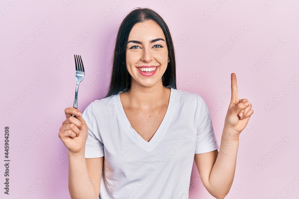 Beautiful woman with blue eyes holding one silver fork smiling with an idea or question pointing finger with happy face, number one Beautiful woman with blue eyes holding one silver fork smiling with an idea or question pointing finger with happy face, number one