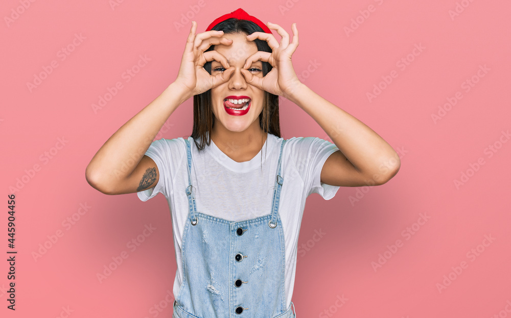 Young hispanic girl wearing casual clothes doing ok gesture like binoculars sticking tongue out, eyes looking through fingers. crazy expression. Young hispanic girl wearing casual clothes doing ok gesture like binoculars sticking tongue out, eyes looking through fingers. crazy expression.