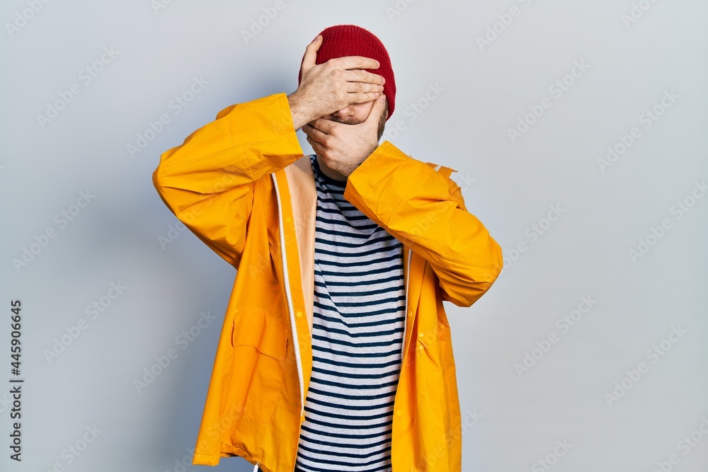 Caucasian man with beard wearing yellow raincoat covering eyes and mouth with hands, surprised and shocked. hiding emotion