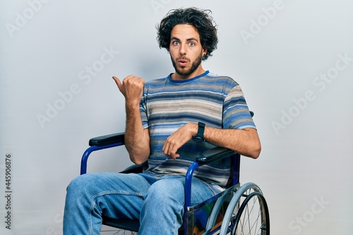 Handsome hispanic man sitting on wheelchair surprised pointing with hand finger to the side, open mouth amazed expression.