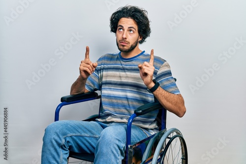 Handsome hispanic man sitting on wheelchair pointing up looking sad and upset, indicating direction with fingers, unhappy and depressed.