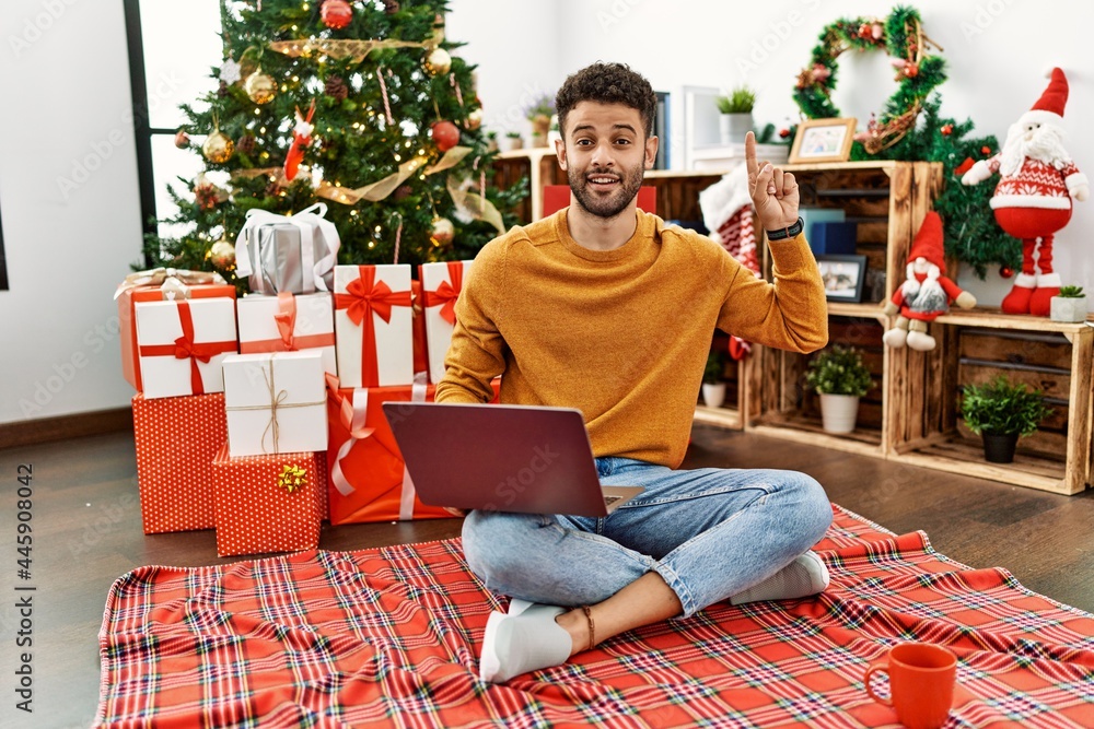 Arab young man using laptop sitting by christmas tree smiling amazed and surprised and pointing up with fingers and raised arms. Arab young man using laptop sitting by christmas tree smiling amazed and surprised and pointing up with fingers and raised arms.
