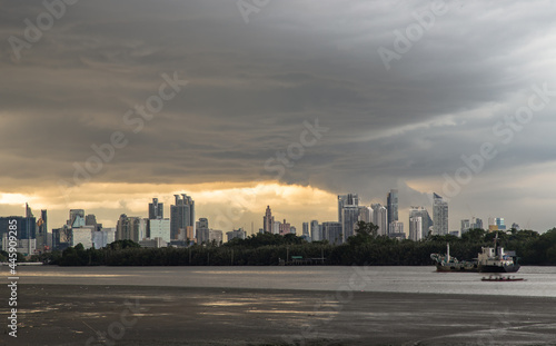 Bangkok, Thailand - 17 Jul 2021 : A cargo ship is sailing in the Chao Phraya River at evening with green forest and skyscrapers at the background. Copy space, Selective focus.