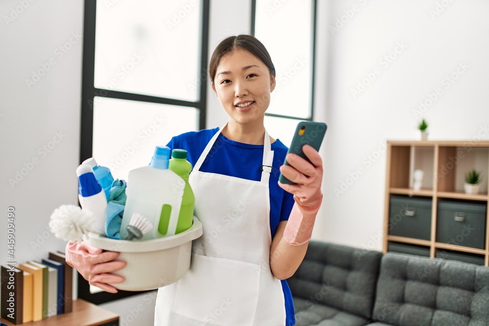 Young chinese housewife holding cleaning products using smartphone at home. Young chinese housewife holding cleaning products using smartphone at home.
