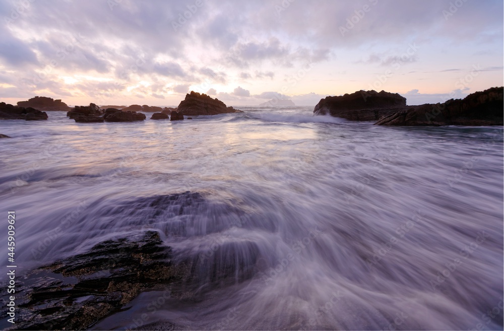 Sunrise scenery of beautiful Wai'ao Beach, in Yilan (Ilan), Taiwan, with turbulent waves rushing upon the rocks & an island on the horizon under moody cloudy sky reflected on sea water (Long Exposure)