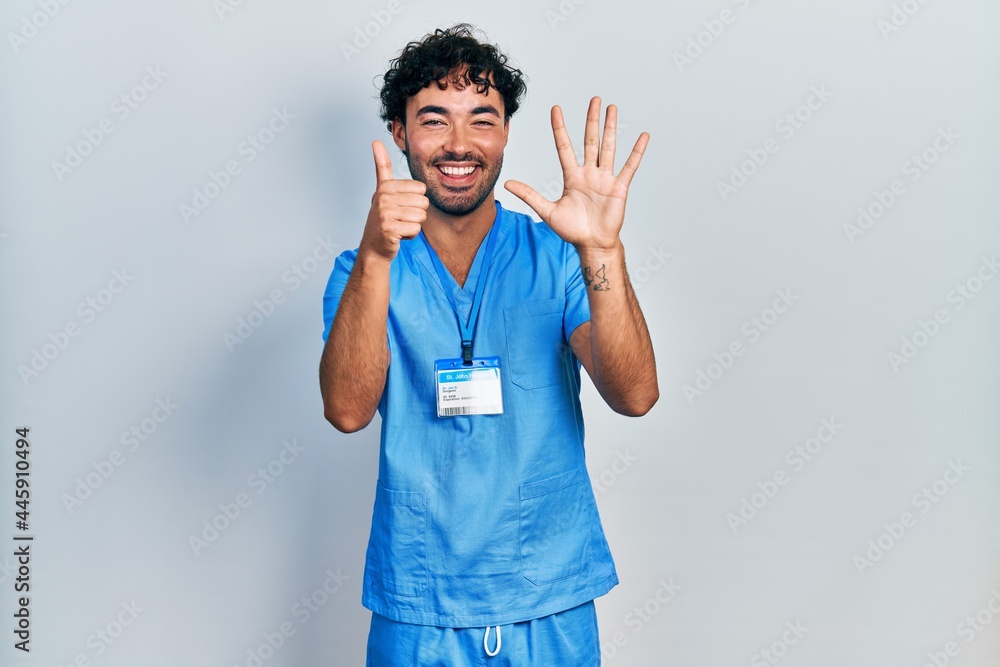 Young hispanic man wearing blue male nurse uniform showing and pointing up with fingers number six while smiling confident and happy. Young hispanic man wearing blue male nurse uniform showing and pointing up with fingers number six while smiling confident and happy.