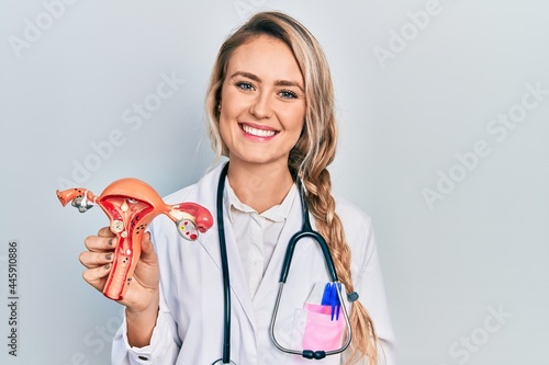 Beautiful young blonde woman holding anatomical model of female genital organ looking positive and happy standing and smiling with a confident smile showing teeth photo