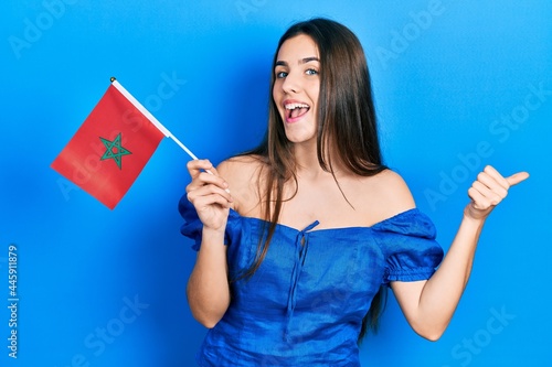 Young brunette teenager holding morocco flag pointing thumb up to the side smiling happy with open mouth