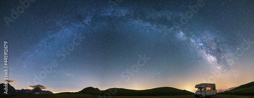 Panoramic night sky over Campo Imperatore highlands, Abruzzo, Italy. The Milky Way galaxy arc and stars over illuminated camper van. Camping freedom in unique hills landscape.