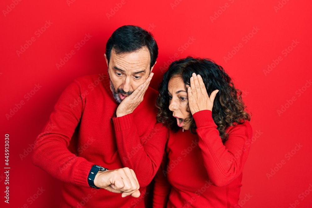 Middle age couple of hispanic woman and man hugging and standing together looking at the watch time worried, afraid of getting late