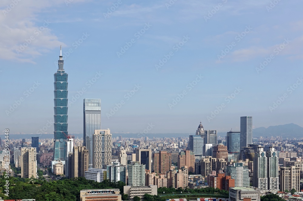 Aerial panorama of Downtown Taipei, vibrant capital city of Taiwan, with prominent Taipei 101 Tower among skyscrapers in Xinyi District & Yangmingshan Mountain on distant horizon under blue sunny sky
