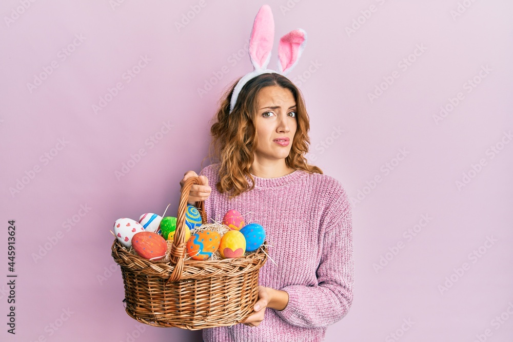 Young caucasian woman wearing cute easter bunny ears holding colored egg clueless and confused expression. doubt concept.