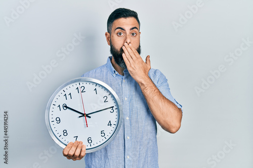 Young hispanic man holding big clock covering mouth with hand, shocked and afraid for mistake. surprised expression