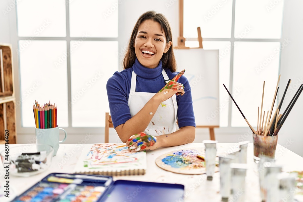 Young brunette woman at art studio with painted hands with a big smile on face, pointing with hand and finger to the side looking at the camera.