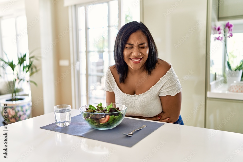 Young hispanic woman eating healthy salad at home with hand on stomach because nausea, painful disease feeling unwell. ache concept. Young hispanic woman eating healthy salad at home with hand on stomach because nausea, painful disease feeling unwell. ache concept.