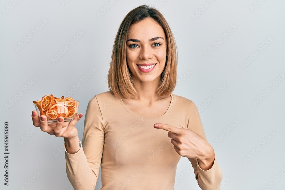 Young caucasian blonde woman holding bowl of dry orange smiling happy pointing with hand and finger
