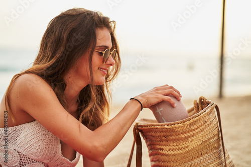 Young hispanic woman taking out a portable speaker from a bag on beach during sunset