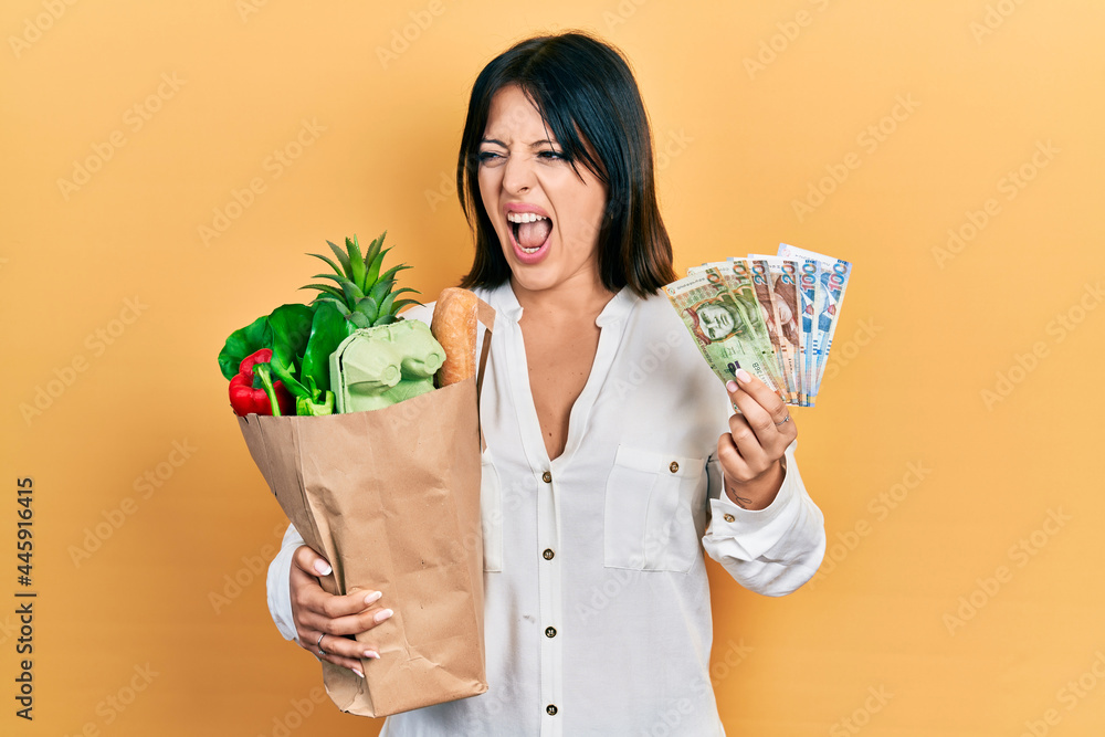 Young hispanic woman holding groceries bag and peruvian sol banknotes angry and mad screaming frustrated and furious, shouting with anger. rage and aggressive concept. Young hispanic woman holding groceries bag and peruvian sol banknotes angry and mad screaming frustrated and furious, shouting with anger. rage and aggressive concept.