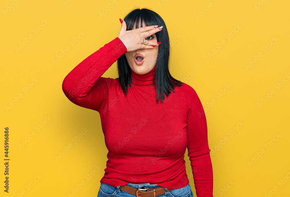 Fototapeta premium Young hispanic woman wearing casual clothes peeking in shock covering face and eyes with hand, looking through fingers with embarrassed expression.