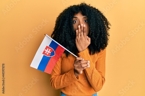 African american woman with afro hair holding slovakia flag covering mouth with hand, shocked and afraid for mistake. surprised expression