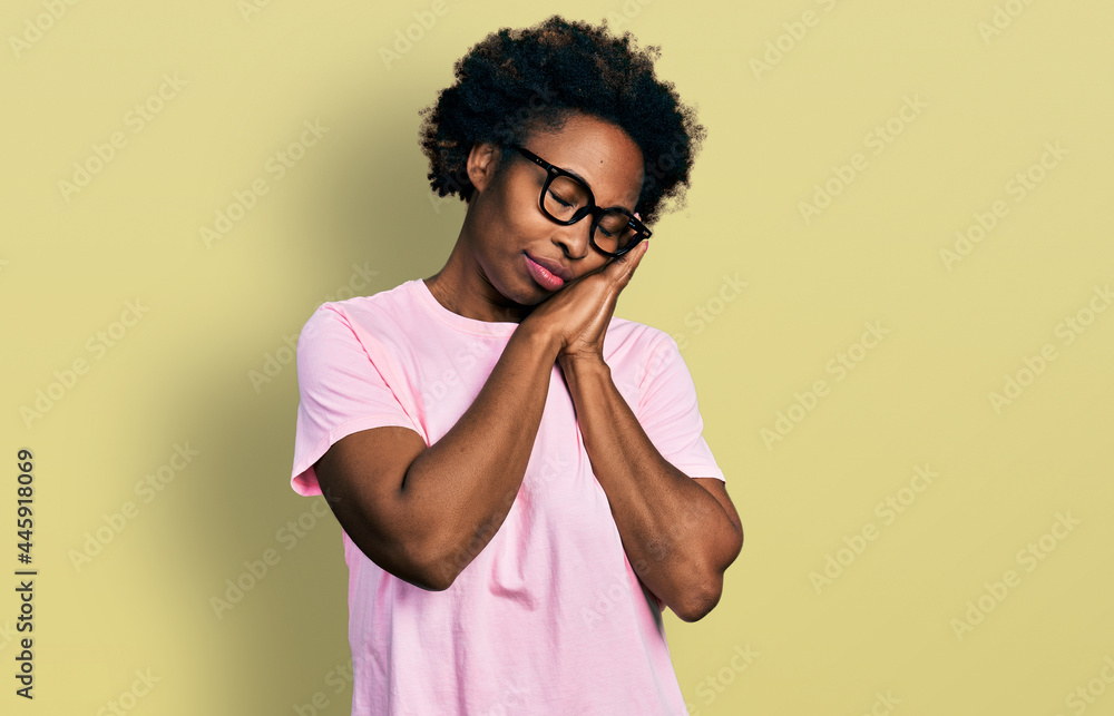 African american woman with afro hair wearing casual clothes and glasses sleeping tired dreaming and posing with hands together while smiling with closed eyes. African american woman with afro hair wearing casual clothes and glasses sleeping tired dreaming and posing with hands together while smiling with closed eyes.