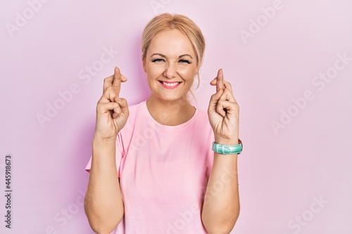 Young blonde woman wearing casual pink t shirt gesturing finger crossed smiling with hope and eyes closed. luck and superstitious concept. © Krakenimages.com