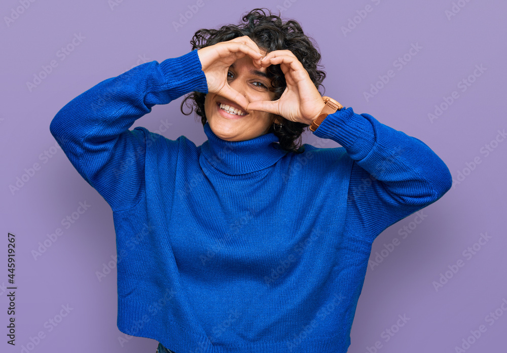 Young hispanic woman with curly hair wearing turtleneck sweater doing heart shape with hand and fingers smiling looking through sign Young hispanic woman with curly hair wearing turtleneck sweater doing heart shape with hand and fingers smiling looking through sign