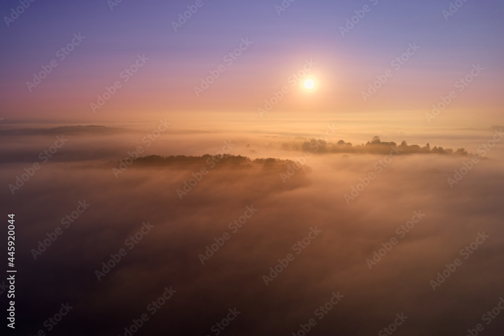 Fog lies over a village. Beautiful landscape after humid night. Sunrise with rich orange and blue colors. Aerial view. Germany, Reudern.