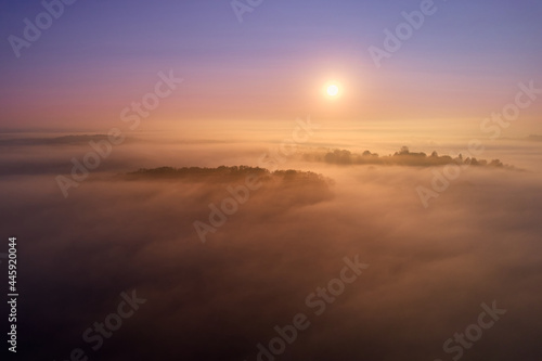 Fog lies over a village. Beautiful landscape after humid night. Sunrise with rich orange and blue colors. Aerial view. Germany, Reudern.