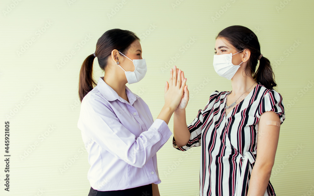 Portrait two Asian and Caucasian young women making high five with confidence, wearing face masks, completely getting vaccination to protect virus with plastic bandage on shoulders, having immunity. Portrait two Asian and Caucasian young women making high five with confidence, wearing face masks, completely getting vaccination to protect virus with plastic bandage on shoulders, having immunity.