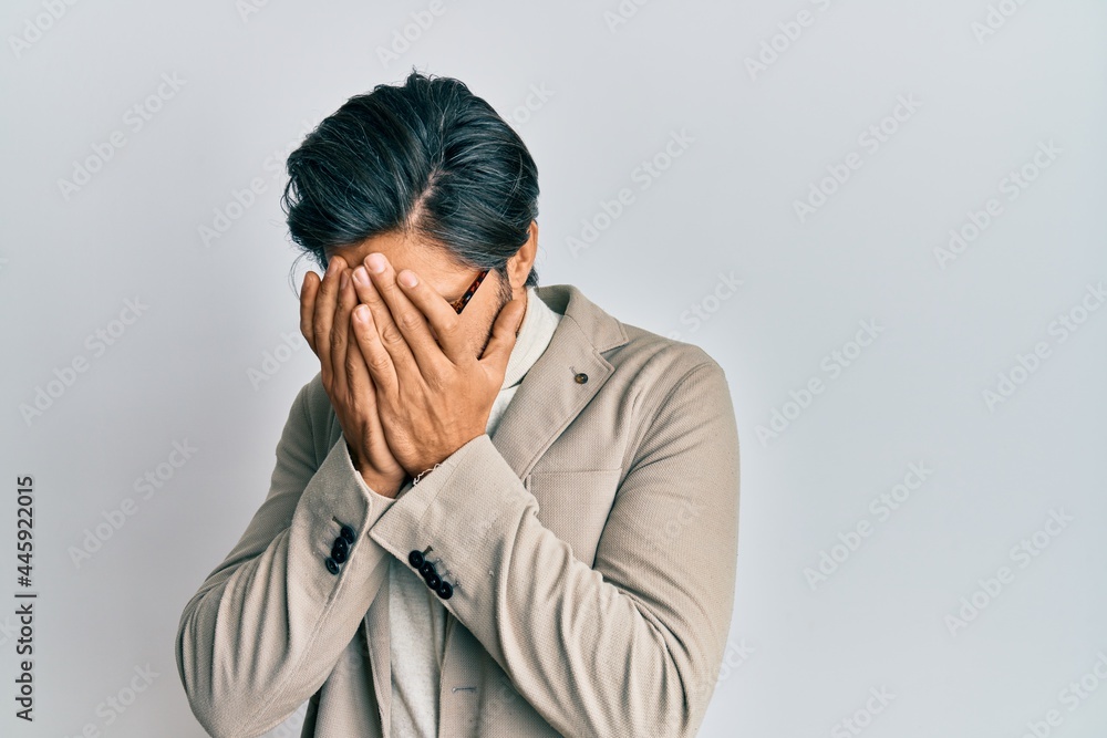Young hispanic man wearing business jacket and glasses with sad expression covering face with hands while crying. depression concept. Young hispanic man wearing business jacket and glasses with sad expression covering face with hands while crying. depression concept.