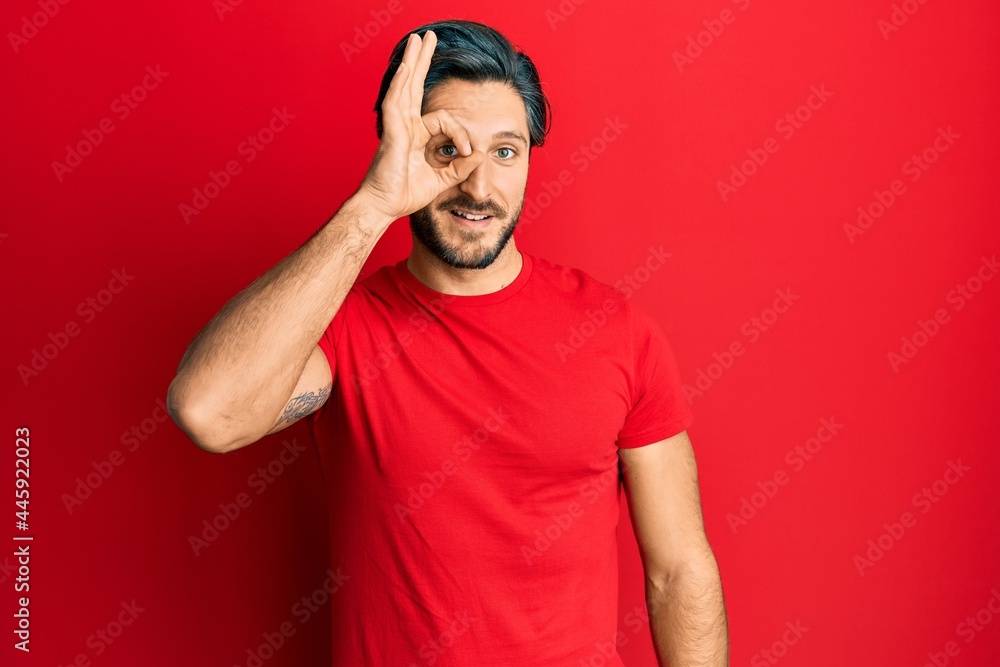 Young hispanic man wearing casual red t shirt doing ok gesture with hand smiling, eye looking through fingers with happy face. Young hispanic man wearing casual red t shirt doing ok gesture with hand smiling, eye looking through fingers with happy face.