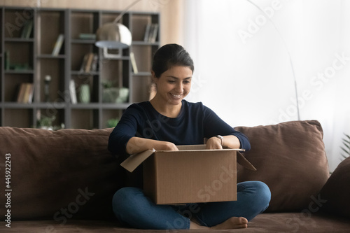 Excited millennial Indian woman sit on sofa at home unpack open box shopping online on internet. Smiling young mixed race female buyer client unbox package with web order. Good delivery concept. © fizkes