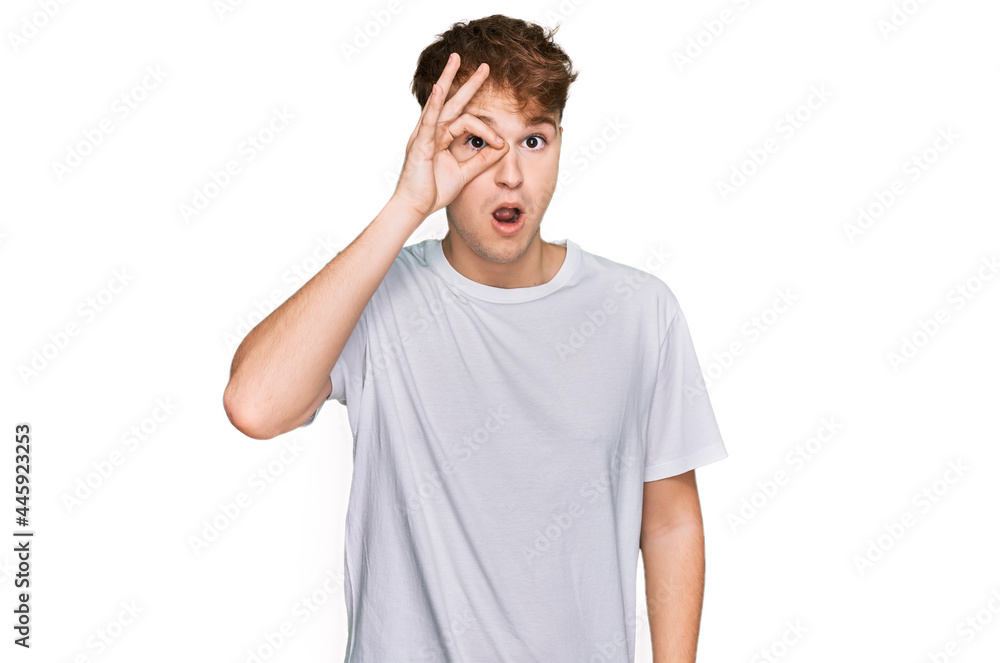 Young caucasian man wearing casual white t shirt doing ok gesture shocked with surprised face, eye looking through fingers. unbelieving expression. Young caucasian man wearing casual white t shirt doing ok gesture shocked with surprised face, eye looking through fingers. unbelieving expression.