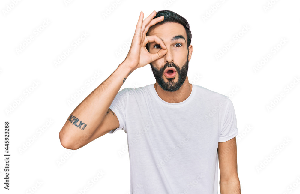 Young hispanic man wearing casual white t shirt doing ok gesture shocked with surprised face, eye looking through fingers. unbelieving expression. Young hispanic man wearing casual white t shirt doing ok gesture shocked with surprised face, eye looking through fingers. unbelieving expression.