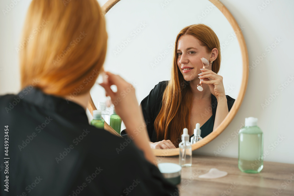 Red hair young girl doing natural beauty routine with gua sha quartz roller at home. Redhead beautiful woman using facial massage stone and looking to the mirror. Beauty routine concept.