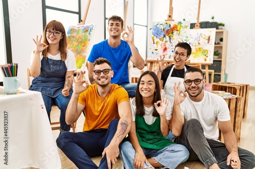 Group of people sitting at art studio doing ok sign with fingers, smiling friendly gesturing excellent symbol