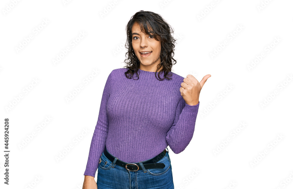 Young hispanic woman wearing casual clothes smiling with happy face looking and pointing to the side with thumb up. Young hispanic woman wearing casual clothes smiling with happy face looking and pointing to the side with thumb up.
