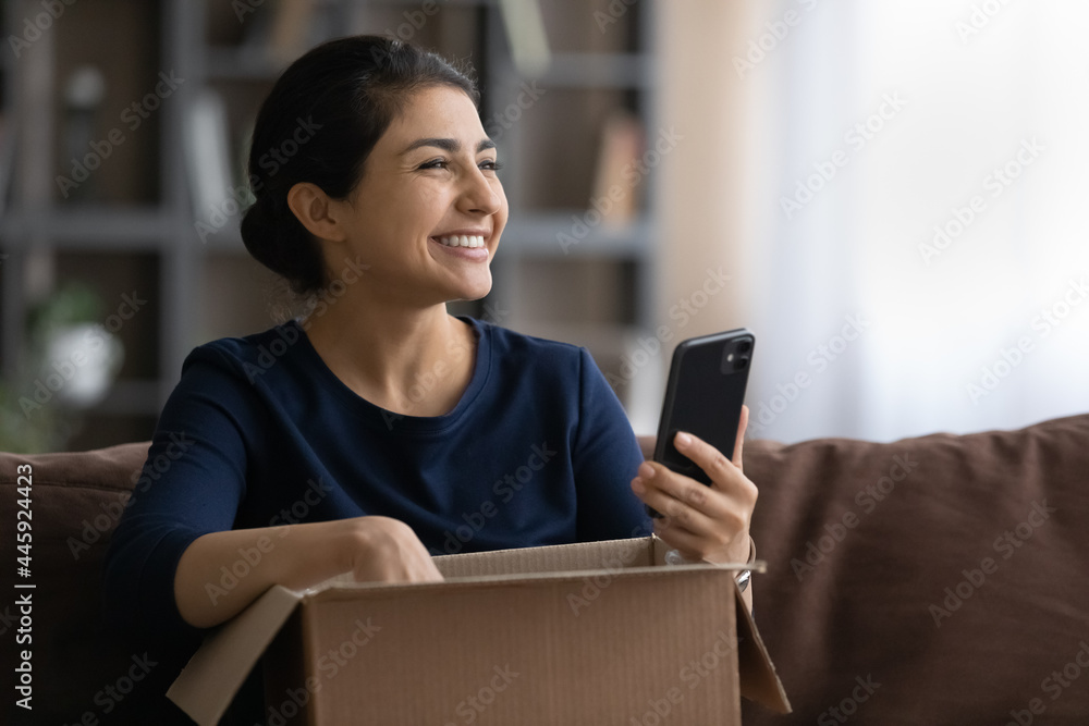 Happy young Indian woman excited open unbox package with internet order track parcel or rate on cellphone. Smiling biracial female buyer unpack box shopping online on smartphone gadget. Happy young Indian woman excited open unbox package with internet order track parcel or rate on cellphone. Smiling biracial female buyer unpack box shopping online on smartphone gadget.