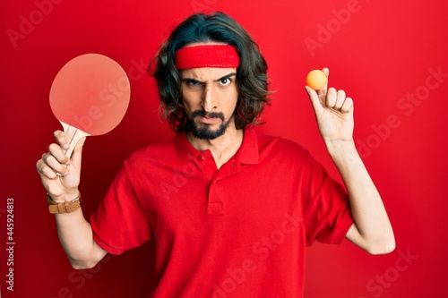 Young hispanic man holding red ping pong racket and ball skeptic and nervous, frowning upset because of problem. negative person.